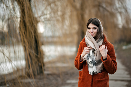 Young attractive brunette girl, with scarf around her neck, in orange coat, looks at camera while standing in autumn park against backdrop of blurry trees and river.