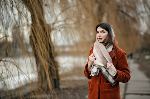 Young attractive brunette girl, with scarf around her neck, in orange coat, holding scarf with her hands, looking away, standing in autumn park against backdrop of blurry trees and river, with copy space.