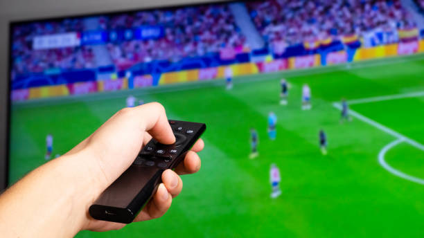 mano dell'uomo che tiene un controller tv nero che guarda una partita di calcio su uno schermo televisivo sullo sfondo - calcio internazionale foto e immagini stock