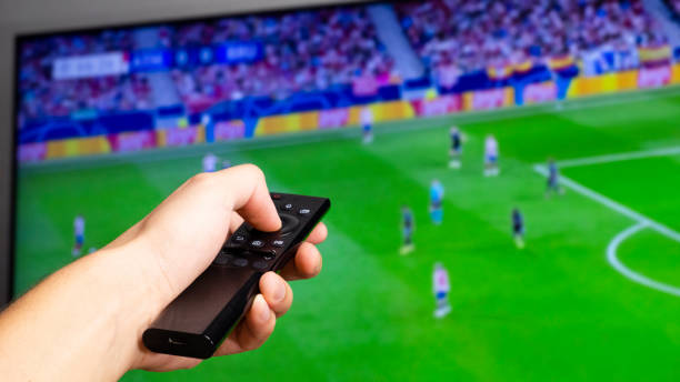 Man hand holding a black tv controller watching a football match on a tv screen in the background Man hand holding a black tv controller watching a football match on a tv screen in the background international team soccer stock pictures, royalty-free photos & images