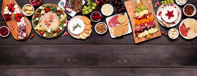 Christmas charcuterie top border against a dark wood banner background. Variety of cheese and meat appetizers. Christmas tree, wreath and candy cane arrangements. Copy space.