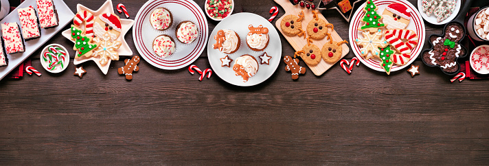 Cute Christmas sweets and cookie top border. Above view on a rustic dark wood banner background with copy space. Fun holiday baking concept.