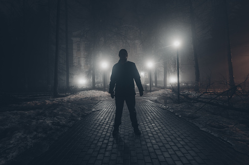 Sad man alone walking along the alley in night foggy park. Back view.