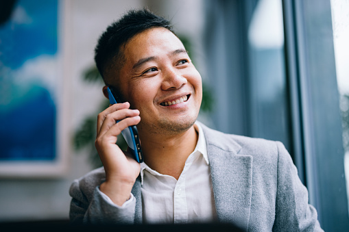 Close up photo of smiling Asian business man speaking on the phone while sitting at restaurant desk with a digital tablet.