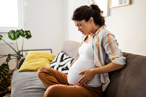 Pregnant young woman grimacing with hands on stomach while sitting at home