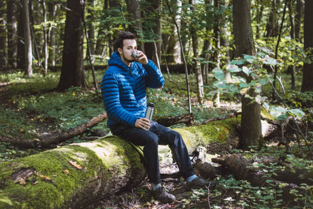 istock Man in hiking clothes rests on a fallen log and drinks hot tea from a thermos. Hiker with blue down jacket. Staying in the fresh air 1437550102