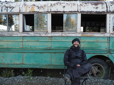 A shot of a smiling Caucasian man sitting in front of an old abandoned bus in Spain