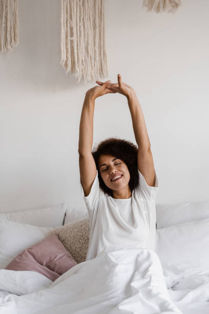 african american woman wake up and enjoying good morning. african girl in pajamas stertching in cozy bed and raising hands up at home. - wake up stretching women black imagens e fotografias de stock