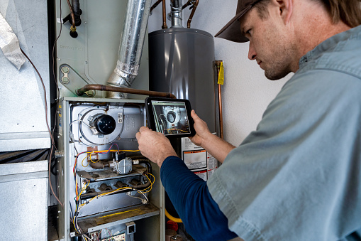 Young Male Property Inspector Photographing a Furnace Hot Water Heater and Air Conditioning Unit Inside a Residential Home Garage