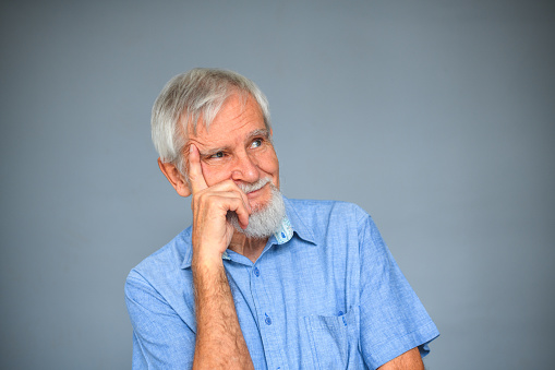 Portrait of elderly man wearing blue shirt, looking up with hand on chin. Thoughtful senior entrepreneur looking for solutions for a problem.