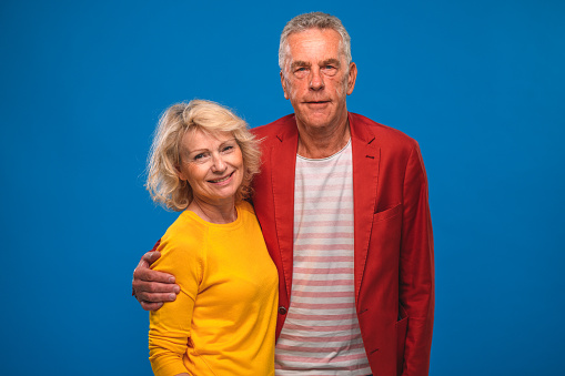 Senior man and mature woman in casual clothing standing with arms around each other. Smiling couple looking at camera and standing against blue background.