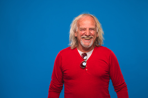 White man with medium-length gray hair and wearing long sleeved red top. Front view of senior man showing joy in blue background.
