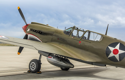 This P-40E Warhawk, also known as a Kittyhawk, wears the U.S. national military insignia of the star inside a blue circle which was superseded on 15 May 1942.   The photo shows a quarter rear profile view of the aircraft in olive drab paint and red spinner, parked with blue sky and clouds behind.  The aircraft wears the shark teeth nose art made famous by the Flying Tigers of the American Volunteer Group (AVG) during WWII.