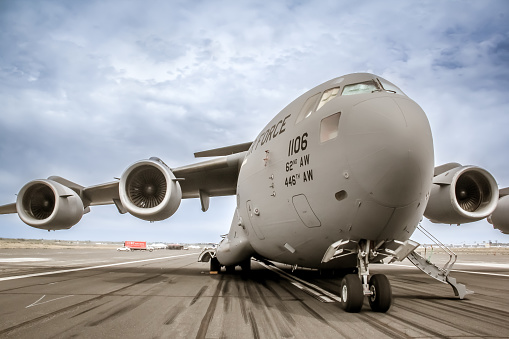 This photo shows a Boeing McDonnell Douglas C-17 jet transport airlifter parked on the ramp.   The C-17 commonly performs tactical and strategic airlift missions by transporting; troops, cargo, medical evacuation, and airdrop duties throughout the world. It is a workhorse of the USAF and are a critical part of the USAF Air Mobility Command's (AMC) mission.  The photo shows a wide angle view of the aircraft from the front right quarter.  The aircraft is painted in Air Force gray with a darkened, cloudy blue sky behind it.