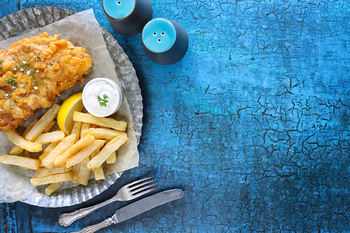 Stock photo showing close-up, elevated view of a greaseproof parchment paper lined plate that is filled with a portion of battered cod and chips, with a lemon slice and tartare sauce.