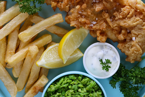 Stock photo showing close-up, elevated view of a portion of battered cod and chips, served with tartare sauce and mushy peas, a lemon slice and parsley garnish on turquoise plate against a yellow background.