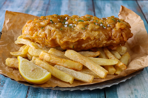 Stock photo showing close-up view of a brown, greaseproof parchment paper lined plate that is filled with a portion of battered cod and chips, with a lemon slice.