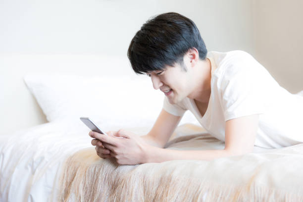 giovane uomo dell'asia orientale che utilizza un telefono mentre è sdraiato sulla schiena su un moderno letto di camera da letto di famiglia - foto stock - smart phone china mobile phone asia foto e immagini stock