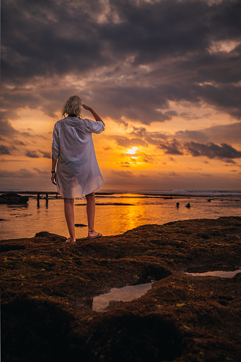 Rear view of a woman in a long white shirt looking  at a dramatic sunset moment, over the Indian Ocean.