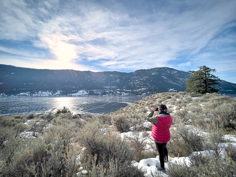 Winter hiking - Eurasian young woman on bluff viewpoint photographing frozen Lake Nicola, Merritt, British Columbia, Canada with smartphone.
