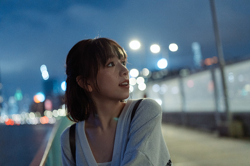 A half-length shot of a young Asian woman looking at the beautiful night views in city of Hong Kong with many neon lights and skyscrapers on the streets.\n\nA portrait of a happy female traveler or tourist feeling amazed by the nightlife of Hong Kong.\n\nA curious Asian girl enjoying the night outdoors near the harbour while looking at the illuminated urban city view of Hong Kong with bright lights.