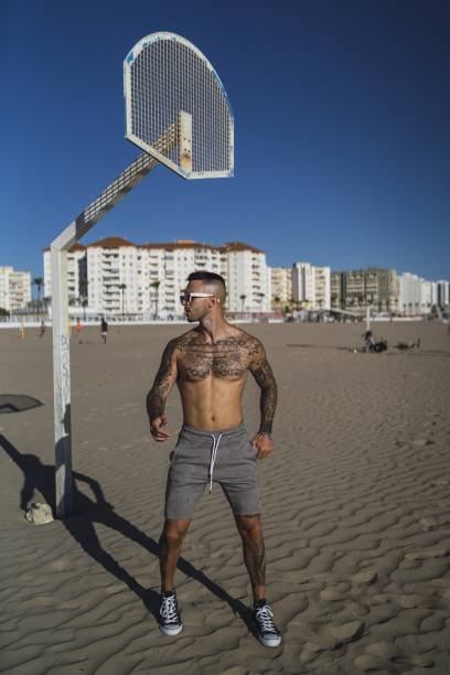 tattoos on his arms, legs, and chest posing on the beach with buildings in the background - fashionable the human body short hair human head imagens e fotografias de stock