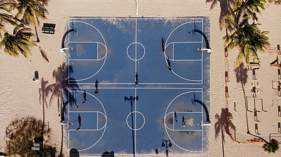 An aerial top view of people playing on blue basketball courts on a sandy beach surrounded by palm trees