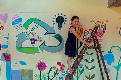 A cute little girl standing on the ladder in front of the mural painted with eco and recycling symbols.