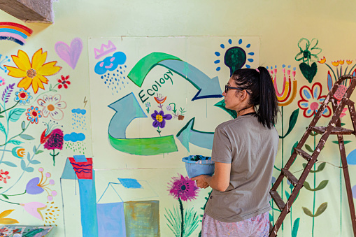 A rear view of one young female artist painting on the wall different symbols of ecology and recycling.