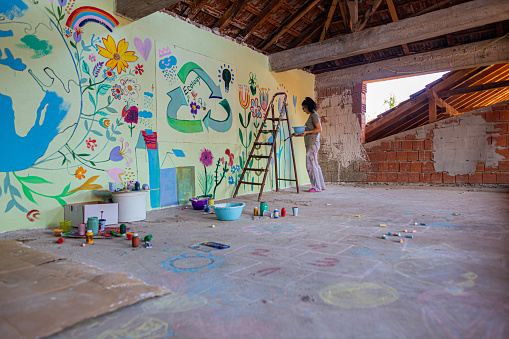 A full-length view of one young female artist painting on the wall different symbols of ecology and recycling.