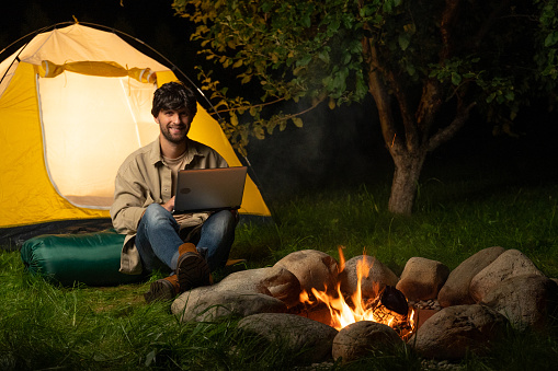 Male traveler sitting by a campfire uses a laptop in a tent camp. A person engaged in digital remote work, electronic purchases, via the Internet.