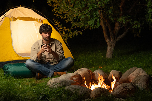 Male tourist settles down next to a tent by the campfire and drinks coffee or tea from a thermos in a pine forest for the weekend