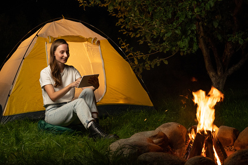 Charming young woman, in a white T-shirt, holds a tablet computer and smiles while resting near a camping tent in the forest by the campfire.