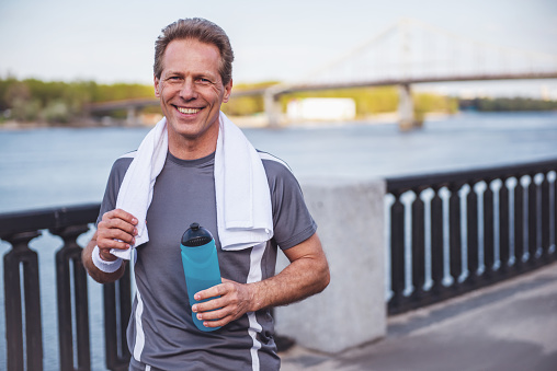 Handsome middle aged man in sports uniform is holding a bottle of water, looking at camera and smiling, resting during morning run
