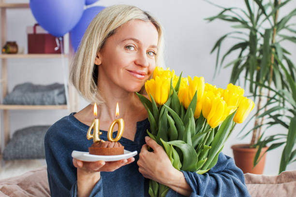 Portrait of happy middle aged woman holding birthday cake with lit candles Smiling Middle aged woman with birthday cake and bouquet of flowers. Adult female celebration her birthday at home woman birthday cake stock pictures, royalty-free photos & images