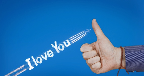 A closeup shot of a person's hand holding a thumbs up with an I love you text made with airplane tracks