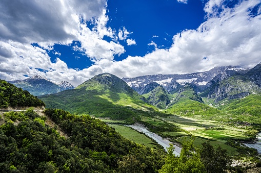 The image shows a view of the Vjosa river valley in Albania with  a beautiful bright cloudy sky and snow capped mountains in the distance