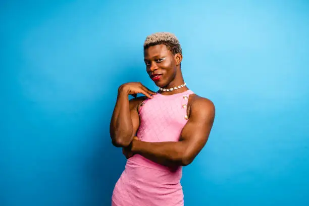 Cheerful African American androgynous person with dyed hair and makeup wearing pink dress standing near blue background and touching shoulder while looking at camera