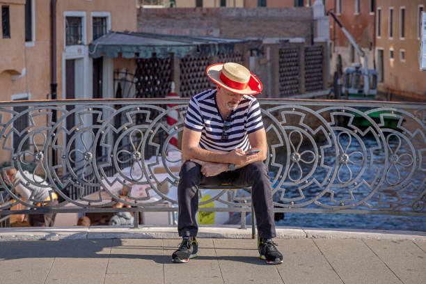 Venice, Italy - October 11th 2022: Male gondolier sitting on a bridge and checking his mobile phone while waiting for customers in the center of the old and famous Italian city Venice