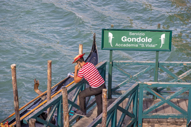 Venice, Italy - October 9th 2022: Male gondolier standing in the sun and checking his phone while he is waiting for customers in the center of the old and famous Italian city Venice