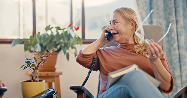 relax, pensionamento e telefonata con una donna anziana su un telefono in un salotto, felice, allegra e parlante. femmina matura che si gode allegra conversazione in rete fissa mentre legge nella sua casa - telefono fisso foto e immagini stock