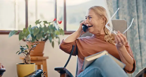 Relax, retirement and phone call with elderly woman on a telephone in a living room, happy, cheerful and talking. Mature female enjoying cheerful landline conversation while reading in her home