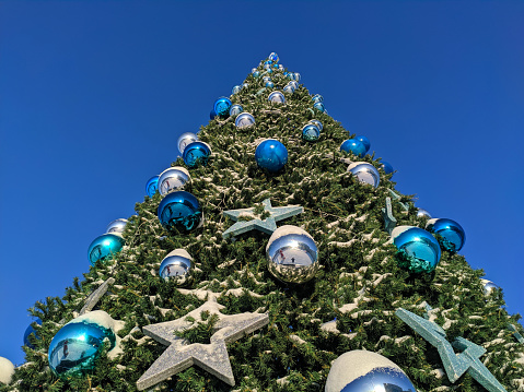 Bottom-up view of a snow-covered Christmas tree on the street decorated with large glass balls and stars.