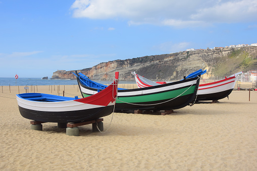 Three traditional brightly painted Portuguese wooden fishing boats sitting on the sand along the beach in Nazare, Portugal with a view of the old town, O Sitio, on top of the cliffs.