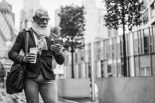 Business hipster senior man using mobile phone while walking to work with city in background - Focus face - Black and white editing