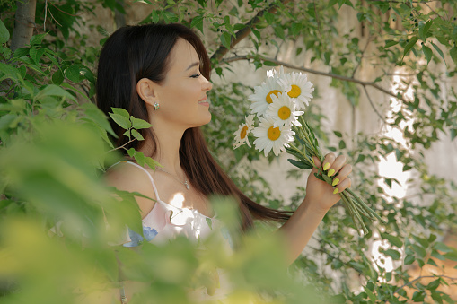 Beautiful young woman stands in the middle of green plants with a bouquet of flowers on a hot summer day