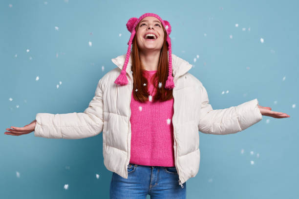 Happy woman in winter coat keeping arms outstretched while snow falling against blue background stock photo