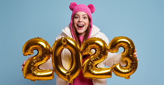 Excited young woman in winter coat holding gold colored numbers against blue background