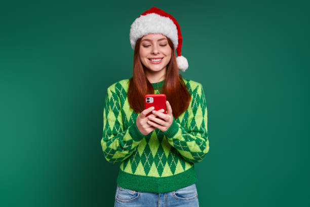Beautiful young woman in Christmas hat using smart phone and smiling against green background stock photo