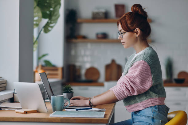 Young redhead woman working on laptop while sitting at the kitchen counter at home stock photo